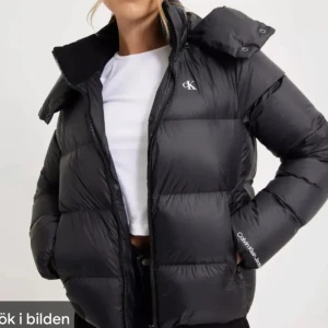 Svart pufferjacka från Calvin Klein - Snygg svart pufferjacka från Calvin Klein med dragkedja och hög krage. Perfekt för kyliga dagar med sin vadderade design. Diskret CK-logga på bröstet och ärmen ger en stilren touch. Säljer då den är lite stor på mig som brukar ha Xs/S 