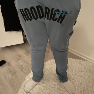 Blå mjukisbyxor från Hoodrich - Säljer ett par blå mjukisbyxor från Hoodrich med stor logga framtill. Byxorna har en bekväm passform och är perfekta för en avslappnad stil. De har elastiska muddar vid bensluten och en praktisk ficka på sidan.