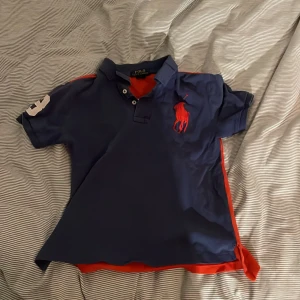 Blå och röd pikétröja från Ralph Lauren - Säljer en snygg blå pikétröja från Ralph Lauren med röd logga på bröstet och nummerdetalj på ärmen. Tröjan har en klassisk krage och knappar framtill. Perfekt för en avslappnad men stilren look!