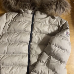 Beige dunjacka från Moncler - Säljer en superfin beige dunjacka från Moncler i mycket bra skick. Jackan har en mysig pälskantad huva och dragkedja framtill. Perfekt för kalla vinterdagar! 🧥❄️