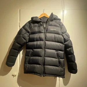 Svart dunjacka från Peak Performance - Säljer en svart dunjacka från Peak Performance i mycket bra skick. Jackan är perfekt för kalla vinterdagar och har en dragkedja framtill. Den är långärmad och har en klassisk pufferstil som håller dig varm och bekväm. Passar både till vardags och utomhusaktiviteter.