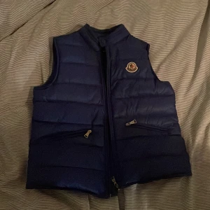 Blå dunväst från Moncler - Säljer en snygg blå dunväst från Moncler i mycket bra skick. Västen har en dragkedja framtill och två praktiska fickor med dragkedjor. Perfekt för höst och vår när du vill hålla dig varm men ändå se stilren ut. Moncler-loggan är broderad på bröstet. Passar både till vardags och lite finare tillfällen.