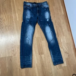 Blå jeans från Supply & Demand - Säljer ett par snygga blå jeans från Supply & Demand i storlek S. De har en cool distressed look med slitningar och färgstänk. Perfekta för en avslappnad stil. Passar bra till både vardag och fest!