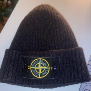  Stone Island mössa - Snygg mössa från Stone Island. Mössan är svart med gul gröna detaljer på badgen. Badgen är fast broderad med knappar på sidorna.Perfekt för att hålla värmen när vintern kommer. Vet ej om den är äkta men ser väldigt fräsch ut. Kan mötas upp eller frakt