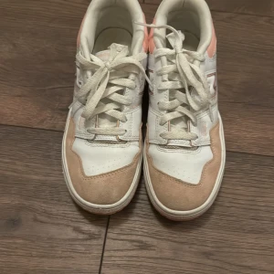 Vita och rosa sneakers från New Balance - Snygga vita sneakers med rosa detaljer från New Balance. Skorna har snörning och en bekväm sula, perfekt för vardagsbruk. Kombinationen av vitt och rosa ger en fräsch look.