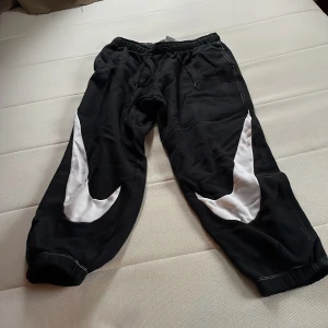 Svarta mjukisbyxor från Nike - Säljer ett par svarta mjukisbyxor från Nike i storlek M. Byxorna har vita detaljer längs sidorna och är perfekta för en avslappnad stil. De är tillverkade i ett mjukt material och har en bekväm passform med resår i midjan.