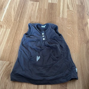Mörkgrå ärmlös klänning från Name It - Söt mörkgrå ärmlös klänning från Name It med tre knappar framtill och en hjärtapplikation på fickan. Perfekt för en avslappnad stil.