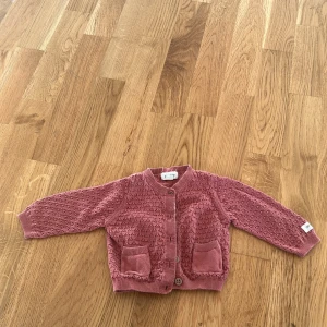 Rosa stickad kofta från Newbie - Söt rosa stickad kofta från Newbie med ett fint mönster och knappar framtill. Perfekt för de små under kyligare dagar. Koftan har långa ärmar och två små fickor fram.