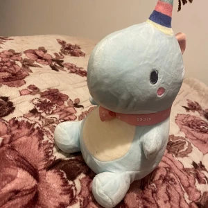 Blå enhörningsmjukisdjur från Momoco - Söt och mjuk enhörning i ljusblått med en färgglad randig horn och rosa detaljer. Den har en rosa rosett runt halsen och små, runda ögon. Perfekt som en kramgo kompis eller dekoration i rummet.