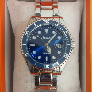 Blå klocka från Comuda - Snygg klocka från Comuda med blå urtavla och datumvisning. Den har en silverfärgad metallarmband och en tydlig kvartsrörelse. Perfekt för både vardag och fest!