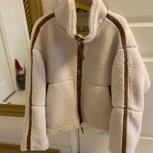 Cremevit teddyjacka med bruna detaljer i storlek M - Supermysig cremevit teddyjacka från H&M med bruna detaljer längs ärmarna och dragkedjan. Perfekt för kyliga höst- och vinterdagar då den är lätt vadderad. Jackan är oversized i storlek. Har bara använt den ett par enstaka gånger. Den är i mycket bra skick. Kan inte se några tecken på användning. Pilejacka. 