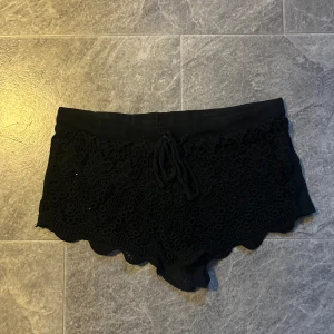 Svarta spets shorts - Snygga svarta shorts med spetsdetaljer och knytning i midjan. Perfekta för en somrig look eller en kväll ute. De har en elegant och feminin design med vackra blommönster i spetsen. AI