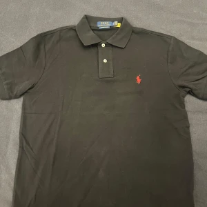 Polo Ralph lauren pike  - Snygg svart pikétröja från Ralph Lauren med röd broderad logga på bröstet. Tröjan har en klassisk krage och knappar framtill. Perfekt för en stilren look. Priser går att diskutera vid snabb affär.