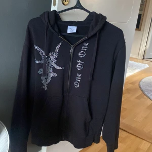 Svart hoodie med paljettdetaljer från One of One - Säljer en svart hoodie från One of One i storlek medium. Den har en cool paljettdekoration av en ängel på framsidan och texten 'one of one'. Perfekt för höst och vinter med sin mysiga känsla och dragkedja framtill. Passar både till vardags och festliga tillfällen! Några av paljetterna har försvunnit som ni ser på bild 2 men inget märkvärdigt. Ord pris 1500kr, pris kan diskuteras😃