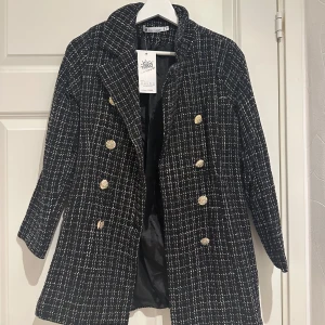 Svartvit rutig kappa från Forest Men - Elegant svartvit rutig kappa från Forest Men med dubbelknäppning och guldknappar. Perfekt för en stilren look under kyligare dagar.
