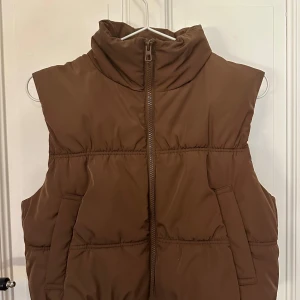 Brun dunväst  - Säljer en brun dunväst från H&M Divided. Västen har en dragkedja framtill och en hög krage för extra värme. Perfekt för kyligare dagar. OBS: Stoppningen i mitten där bak har ”flyttat” på sig, det är inget man ser utan endast om man känner med handen. Men den är fortfarande varm ☺️
