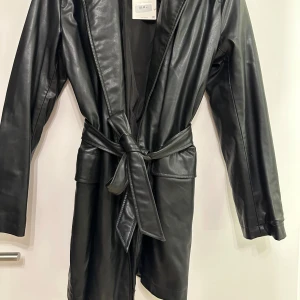 Svart skinnkavaj/klänning från Zara - Snygg svart skinnjacka från Zara med knytbälte i midjan. Jackan har en klassisk krage och långa ärmar, perfekt för en stilren look. Går också att använda som en klänning. Aldrig använd säljs för 300kr 