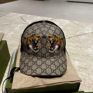 Gucci keps med tigermotiv - Säljer en snygg Gucci keps med deras ikoniska mönster och coolt tigerbroderi framtill. Kepsen har en justerbar rem baktill för perfekt passform. Perfekt för att ge din outfit en lyxig touch!