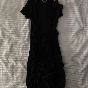 Svart klänning med rynkdetaljer - Snygg svart klänning med rynkdetaljer längs sidorna som ger en smickrande passform. Klänningen har korta ärmar och är tillverkad i ett mjukt material som ger en bekväm känsla. Perfekt för en stilren look. ⚠️ni betalar för frakt⚠️