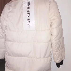 Vit pufferjacka från Calvin Klein Jeans - Säljer en snygg vit pufferjacka från Calvin Klein Jeans. Jackan har en dragkedja framtill och praktiska fickor med dragkedjor. Perfekt för kalla dagar och ger en stilren look. Stor logga på ryggen för extra stilpoäng!