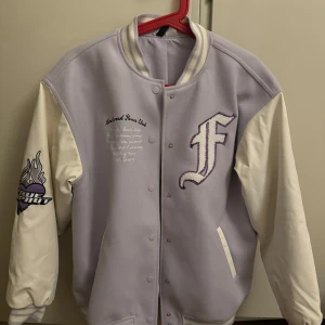 Ljuslila bomberjacka från H&M Divided - Snygg ljuslila bomberjacka från H&M Divided med vita ärmar och broderad detalj på bröstet. Jackan har knappar framtill och ribbade muddar vid ärmslut och krage. Perfekt för en avslappnad stil.