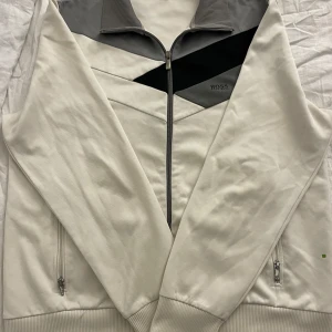 Vit jacka från Hugo Boss - Snygg vit jacka från Hugo Boss med dragkedja och grå detaljer. Jackan har en stilren design med ribbade muddar och två fickor med dragkedja. Perfekt för en casual look.