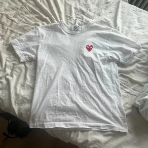 Vit t-shirt från Comme des Garçons - Säljer en vit t-shirt från Comme des Garçons med det ikoniska röda hjärtat på bröstet. T-shirten är i bomull och har en snygg passform. Perfekt för en stilren och casual look. Använd den till vardags eller när du vill ha en enkel men snygg outfit. Storlek m men passar s