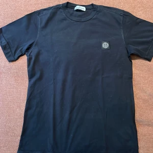 Svart t-shirt från Stone Island Junior - Säljer en stilren svart t-shirt från Stone Island Junior. Den har en klassisk rund hals och korta ärmar. På bröstet finns det ikoniska Stone Island-märket. Perfekt för en casual look eller att styla upp med en jacka. 