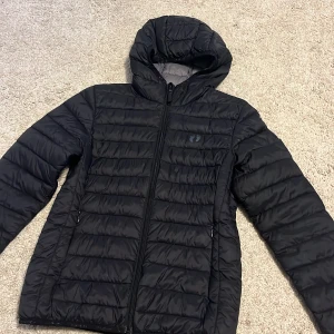Svart pufferjacka med huva - Säljer en svart pufferjacka i mycket bra skick. Den har en praktisk huva och dragkedja framtill. Jackan är perfekt för kyliga dagar och har långa ärmar med elastiska muddar. Den quiltade designen ger extra värme och stil. Perfekt för både höst och vinter!