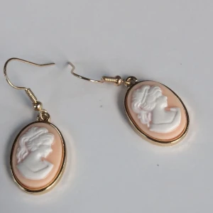 Vintageinspirerade cameo-örhängen - Eleganta vintageinspirerade cameo-örhängen med en klassisk design. De har en vacker vit profil av en kvinna mot en ljusrosa bakgrund, inramad i en gyllene oval.NYA Material, märke ospecificerat postar varje fredag, söndag 