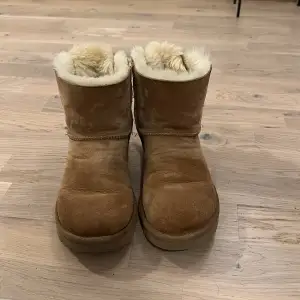 Säljer ett par mysiga bruna UGG boots i mocka med fluffigt foder och söta rosetter bak. Perfekta för kalla dagar när du vill hålla fötterna varma och bekväma. Sulan är robust och ger bra grepp. Använd dem till vardags eller när du vill ha en avslappnad stil.