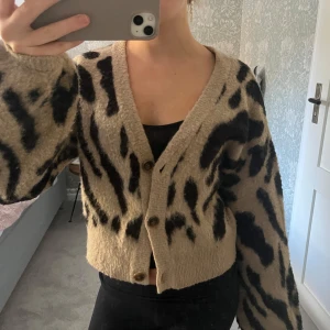Beige och svart kofta med leopardmönster - Säljer en supermysig beige kofta med svart leopardmönster. Den har en v-ringad design och knappar framtill. Perfekt för höst och vinter. Har ett litet hål i armen annars super skick!