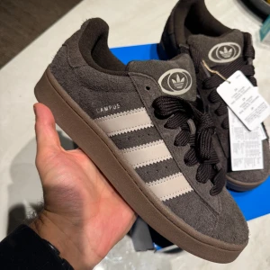 Adidas Campus 00s  - Adidas Campus 00s i färgen dark brown and grey. Nya i kartong med etiketterna kvar.   Storlek: 41 1/3 Unisex  Hämtas i Hammarby Sjöstad alternativt fraktas på köparens bekostnad.