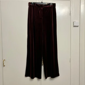 Vinröda sammetbyxor från NA-KD - Säljer ett par superchica vinröda byxor i sammet från NA-KD. De är högmidjade och har en loose fit med vida ben, perfekt för en festlig kväll eller en mysig höstlook. Byxorna har ett knytband i midjan för extra stilpoäng. Helt nya med tag kvar!