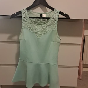 Mintgrön topp med spetsdetaljer från Divided - Säljer en superfin mintgrön topp från Divided i storlek S. Den har vackra spetsdetaljer upptill och är ärmlös med en peplumformad nederdel. Perfekt för sommarens fester eller en dag på stan! 💚