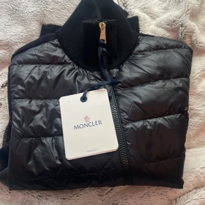 Svart Cardigan från Moncler - Säljer en snygg svart cardigan från Moncler. Jackan har en quiltad design med dragkedja och långa ärmar. Perfekt för kyliga dagar och ger en stilren look. Märkesloggan syns på ärmen. Passar både till vardags och lite finare tillfällen. Helt oanvänd och kvitto, påse och kvitto finns kvar.