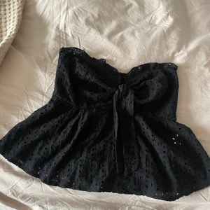 Svart axelbandslös topp med knyt - Säljer en superfin svart axelbandslös topp med knyt framtill. Den har ett sött broderat mönster och är perfekt för sommar eller fest. 