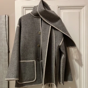 Grå broderad jacka med scarf från Totême - Säljer en elegant grå scarfjacka från Totême med och fina broderade detaljer. Jackan har en normal passform och är perfekt för höst och vår. Den är långärmad och har knappar framtill. Superfin och stilren design!
