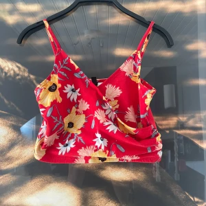 Röd blommig omlottopp - Säljer en superfin röd omlottopp med blommigt mönster i gult och vitt. Toppen har smala axelband och en omlottdesign som ger en snygg passform. Perfekt för sommardagar eller en festlig kväll! 🌸