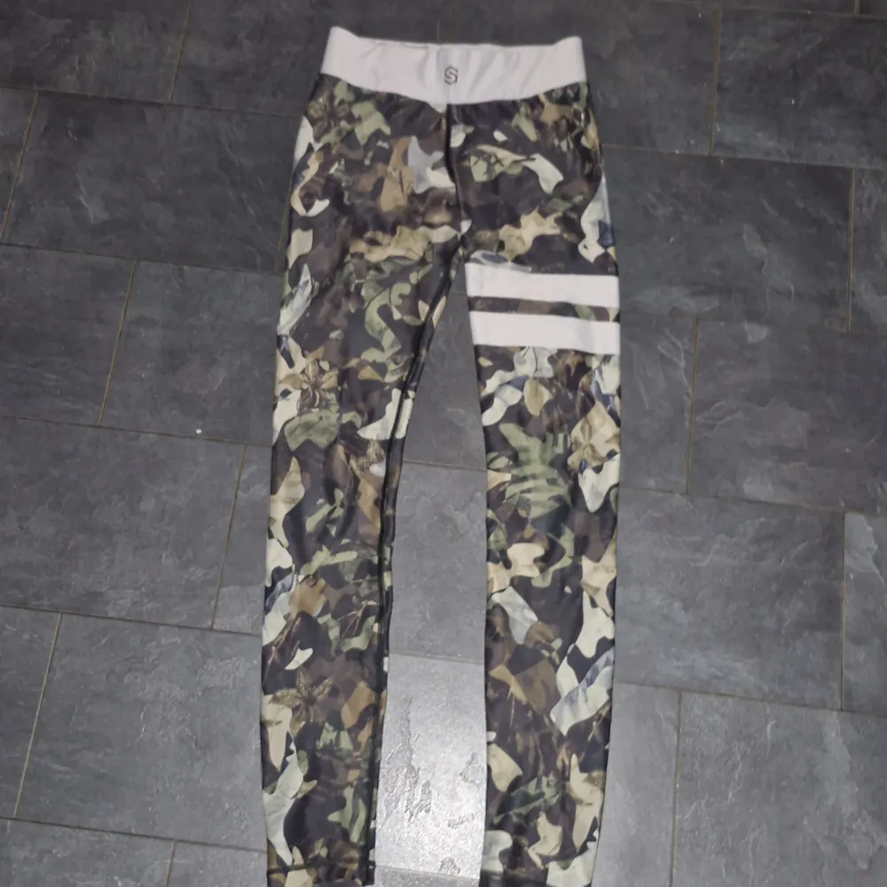 Säljer ett par coola camo leggings med vita detaljer. De är tighta och perfekta för träning eller en avslappnad dag. Mönstret är i olika gröna och bruna nyanser, vilket ger en snygg militärlook. De har en stretchig midja för extra komfort. Passar perfekt för dig som vill ha en stilren och bekväm outfit!. Jeans & Byxor.