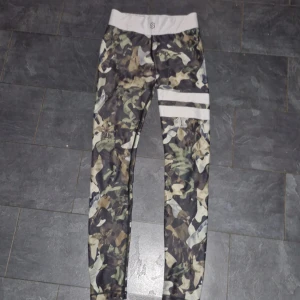 Camo leggings med vita detaljer - Säljer ett par coola camo leggings med vita detaljer. De är tighta och perfekta för träning eller en avslappnad dag. Mönstret är i olika gröna och bruna nyanser, vilket ger en snygg militärlook. De har en stretchig midja för extra komfort. Passar perfekt för dig som vill ha en stilren och bekväm outfit!