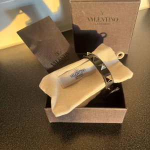 Svart armband med nitar från Valentino - Snyggt svart armband från Valentino med coola silverfärgade nitar. Perfekt för att ge din outfit en edgy touch. Armbandet kommer i originalförpackning och är i mycket bra skick. Passar både till vardags och fest!
