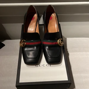 Svarta loafers med klack från Gucci - Säljer ett par eleganta svarta loafers från Gucci, model Marmont i skinn med en stilren mellanhög klack (7 cm)  Skorna har det ikoniska gröna och röda bandet med Guccis klassiska dubbel G framtill. Perfekta för både vardag och fest! 🖤