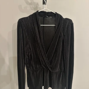 Svart omlottopp från Soaked in Luxury - Snygg svart omlottopp från Soaked in Luxury i ett ribbat material. Perfekt för både vardag och fest! Den har långa ärmar och en elegant omlottdesign som ger en stilren look. Passar bra till både jeans och kjol. 🖤