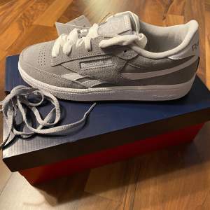 Säljer ett par grå Reebok Club C Revenge Kids sneakers som är helt nya, aldrig använda. Skorna har en klassisk design med vit sula och snörning. Perfekta för både vardag och sportiga aktiviteter. De är tillverkade i syntetmaterial och har en snygg gummisula för bra grepp. Säljer då de var för stora för mig. 