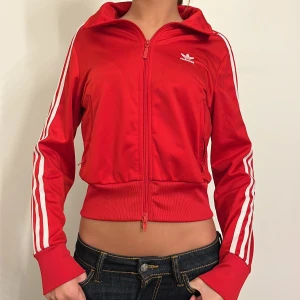 Röd träningsjacka från Adidas - Säljer en snygg röd träningsjacka från Adidas med vita ränder längs ärmarna. Jackan har dragkedja och är i mycket bra skick. Perfekt för träning eller som en sportig vardagslook. Tillverkad med Parley Ocean Plastic, vilket gör den både stilren och miljövänlig. Passar perfekt för höst och vår!