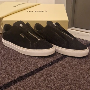 Svarta sneakers från Axel Arigato - Säljer ett par stilrena svarta sneakers från Axel Arigato i mocka. De har en snygg dragkedja framtill och en vit sula som ger en fräsch kontrast. Perfekta för både vardag och fest! Skorna är i bra skick och kommer med originalförpackning.
