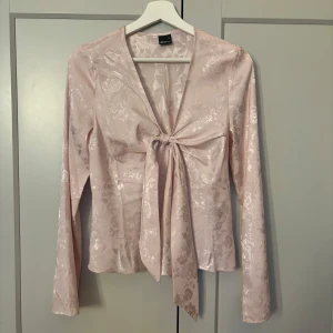 Ljusrosa blus med knytning! - Säljer en elegant ljusrosa blus med knyt framtill. Blusen har ett subtilt blommönster och långa ärmar, perfekt för både vardag och fest. Materialet känns lyxigt och ger en fin glans. Passar perfekt till både jeans och kjol!storlek 36, men passar som xs/s💕
