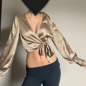 Beige omlottopp i satin - Snygg beige omlottopp i satin med långa ärmar och knytning i midjan. Perfekt för en festlig kväll eller en stilren vardagslook. Toppen har en v-ringad design som ger en elegant touch. Passar bra till både jeans och kjol!