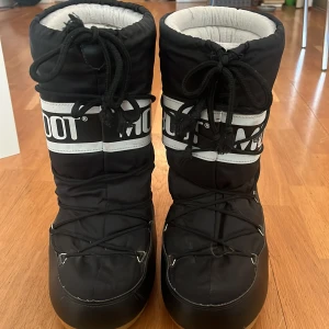 Svarta Moon Boots - Säljer ett par svarta Moon Boots i begagnat skick som anges på bilderna, en sula har gått sönder men är lagad. De är perfekta för vintern med sin varma och vadderade insida. Skorna har snörning framtill och en robust sula som ger bra grepp på isiga underlag. De är både snygga och praktiska för kalla dagar. PRIS KAN DISKUTERAS